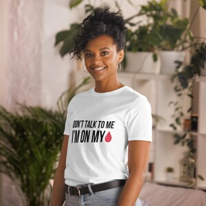 Könnte beinhalten: Weißes T-Shirt mit schwarzem Text, der "Sprich mich nicht an, ich habe meine Periode" lautet, mit einem roten Tropfen-Symbol.