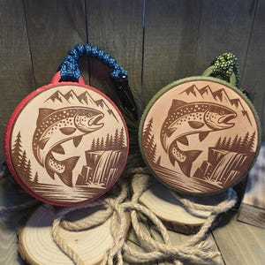 May include: Two round wooden ornaments with a fish and mountain scene carved into them. One has a red border and blue cord, the other has a green border and green cord. Both have a black clip.