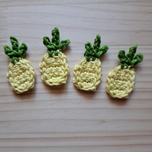 May include: Four small, crocheted pineapple appliques. The pineapples are yellow with green tops, and are arranged in a row. The appliques are handmade and have a textured appearance, suggesting a craft project or DIY item.