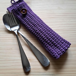May include: A purple crocheted utensil pouch featuring a small loop at the top and a single dark brown wooden button closure. A stainless steel fork and a stainless steel spoon are positioned diagonally across the front of the fabric pouch.