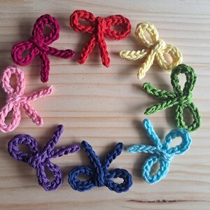 May include: Eight crocheted scissor-shaped decorations in a circular arrangement. The scissors are in various colours including pink, red, yellow, green, blue, and purple. They are set against a light brown wooden background.