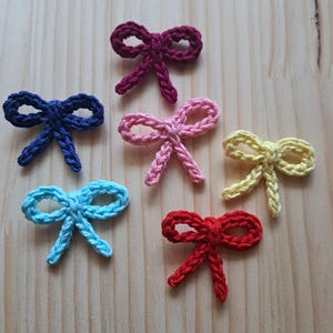 May include: A collection of six crocheted bows in various colors, including blue, pink, yellow, red, and purple. The bows are arranged on a light wooden surface, showcasing their handmade texture and vibrant hues.