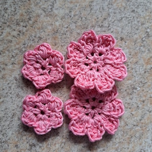 以下が含まれることがあります： さまざまなサイズの4つの手作りのピンク色の手編みの花が、テクスチャ加工された表面に配置されています。 花は、糸の質感を際立たせる、花びらのようなエッジを持つ、詳細なオープンセンターデザインが特徴です。 クラフトプロジェクトに適しています。