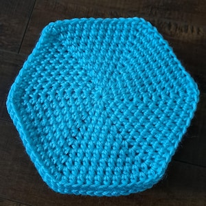 May include: A handmade, turquoise blue crochet hexagon. The crocheted item is made with a tight stitch pattern, and the edges are well-defined. The item is sitting on a dark brown wooden surface.