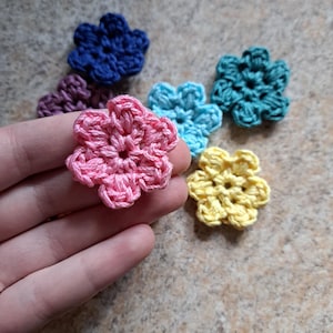May include: A selection of small, handmade crochet flowers in shades of pink, blue, teal, yellow, and purple. Each flower has a five-petal design and a central hole. These colourful flowers are suitable for various craft projects.
