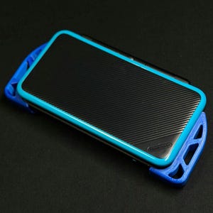 Puede incluir: Una consola de juegos portátil negra con un marco y detalles azules. El dispositivo tiene una superficie texturizada y está colocado en un soporte azul. El dispositivo está sobre una superficie negra.