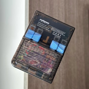 Puede incluir: Una consola de juegos portátil transparente con botones azules. La consola tiene el texto "Miyoo-mini" y especificaciones técnicas impresas. La pantalla muestra un juego colorido. El dispositivo está fabricado en China.