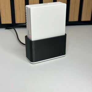 May include: A white electronic device is docked in a black charging base. The device has a rectangular shape with a smooth surface. The charging base has a minimalist design with a white trim. A black cable extends from the base.