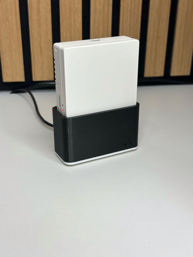 May include: A white electronic device is docked in a black charging base. The device has a rectangular shape with a smooth surface. The charging base has a minimalist design with a white trim. A black cable extends from the base.