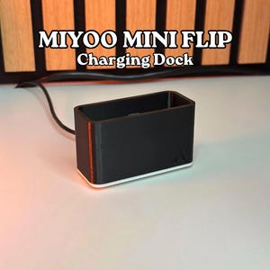 May include: A black charging dock with the text "MIYOO MINI FLIP Charging Dock" in white. The dock has a rectangular shape with a red stripe on the side and a white base. A black cable extends from the dock.