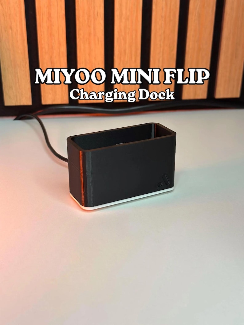 May include: A black charging dock with the text "MIYOO MINI FLIP Charging Dock" in white. The dock has a rectangular shape with a red stripe on the side and a white base. A black cable extends from the dock.