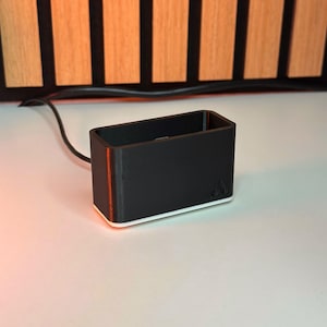 May include: A black rectangular object with a white base and a thin orange stripe on the side. The object has a small logo on the side and is connected to a black wire. The background features vertical wooden slats.