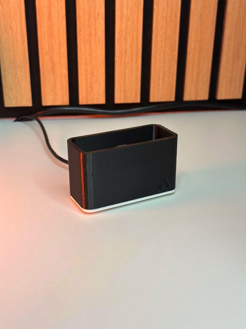 May include: A black rectangular object with a white base and a thin orange stripe on the side. The object has a small logo on the side and is connected to a black wire. The background features vertical wooden slats.