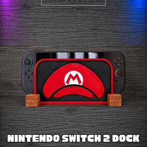 Può includere: Una cover per dock Nintendo Switch 2 nera con un emblema rosso a forma di cappello di Mario e un logo M bianco. La cover è sostenuta da due piccoli blocchi marroni con motivo a mattoni. Il testo AMONEE e NINTENDO SWITCH 2 DOCK COVER sono presenti.