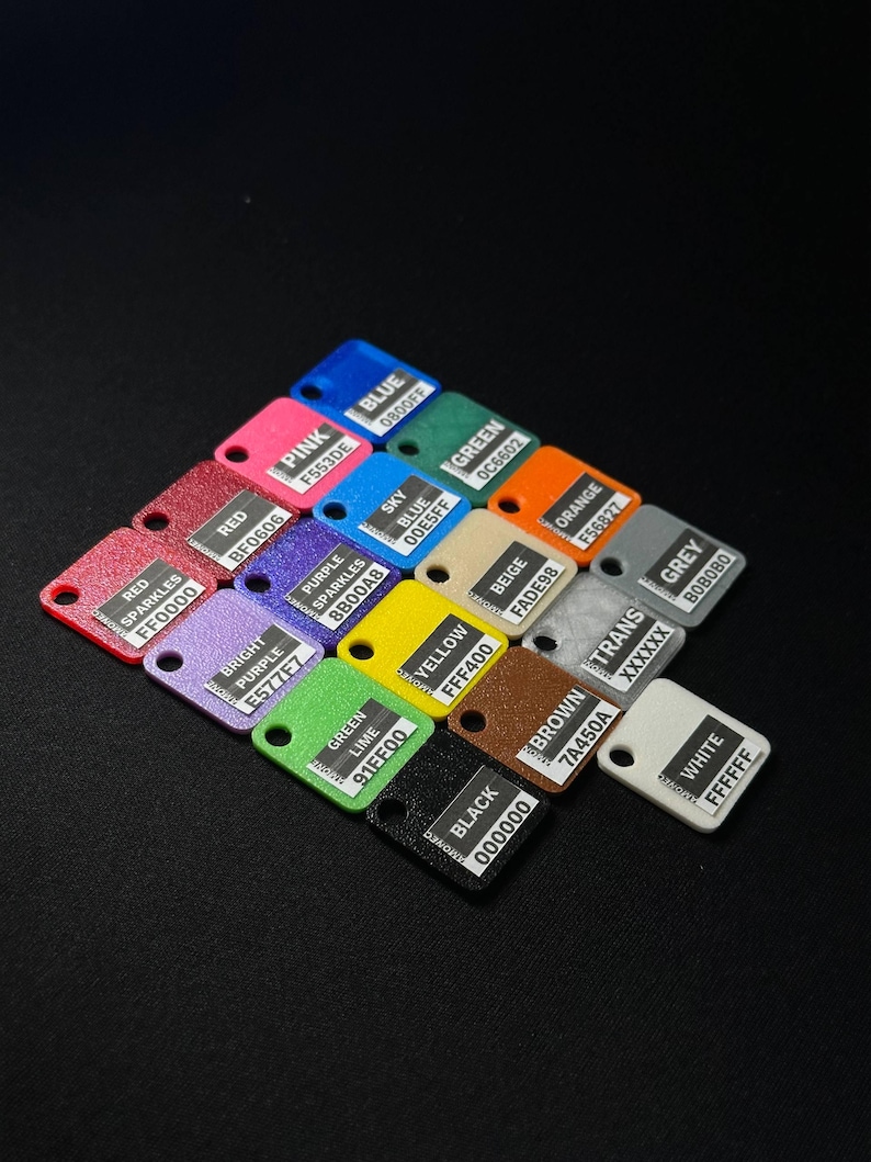 May include: A collection of small, color-coded rectangular tags with rounded corners, each displaying a different color name and hexadecimal code. Colors include red, pink, blue, green, yellow, and black. The tags are arranged on a black surface.
