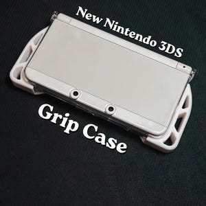 May include: A white New Nintendo 3DS handheld game console with a white grip case. The text "New Nintendo 3DS" and "Grip Case" are printed on the image. The console is on a dark green surface.