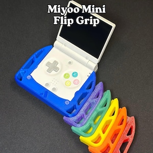 Può includere: Console di gioco portatile Miyoo Mini bianca e blu con schermo nero, abbinata a un set di Flip Grips colorati. Le impugnature sono nei colori dell'arcobaleno: rosso, arancione, giallo, verde, viola.