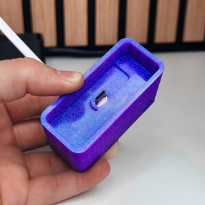 May include: A rectangular, purple and blue resin charging dock with a USB-C port. The dock is held in a hand and has a glittery, iridescent finish. The background is a wooden panel.