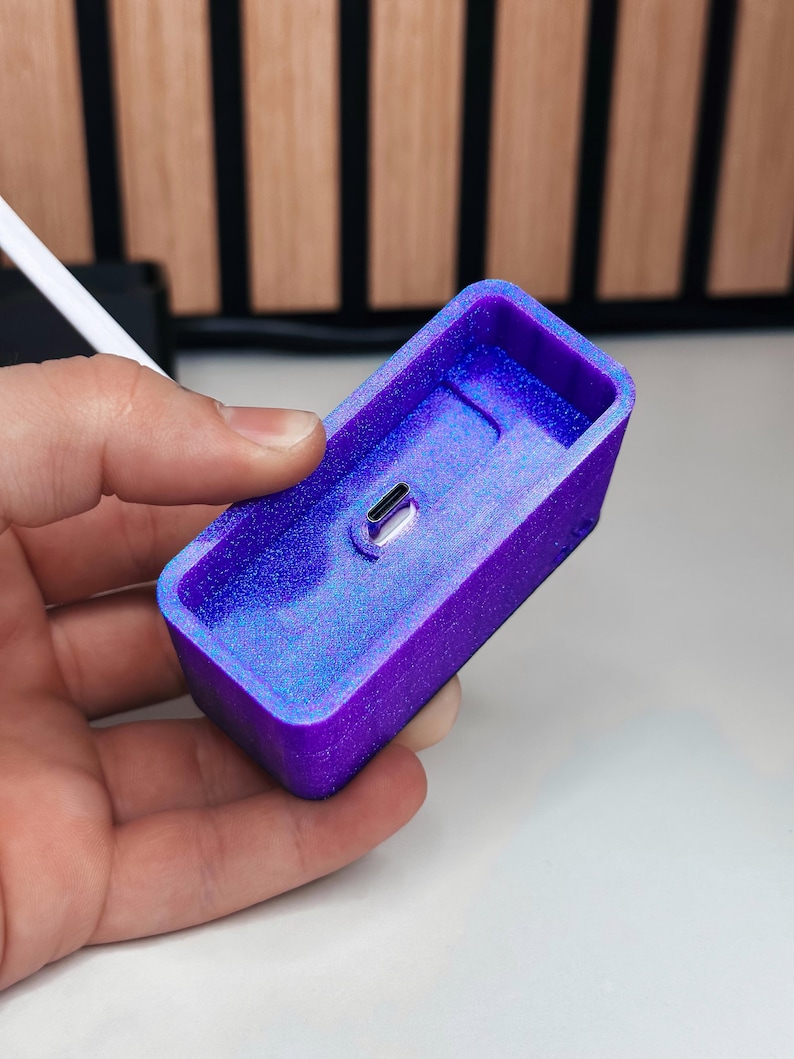 May include: A rectangular, purple and blue resin charging dock with a USB-C port. The dock is held in a hand and has a glittery, iridescent finish. The background is a wooden panel.