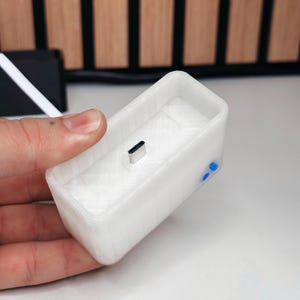 May include: A white rectangular charging dock with a USB-C port. The dock has a textured surface and blue accents on the side. The item is held in a hand.