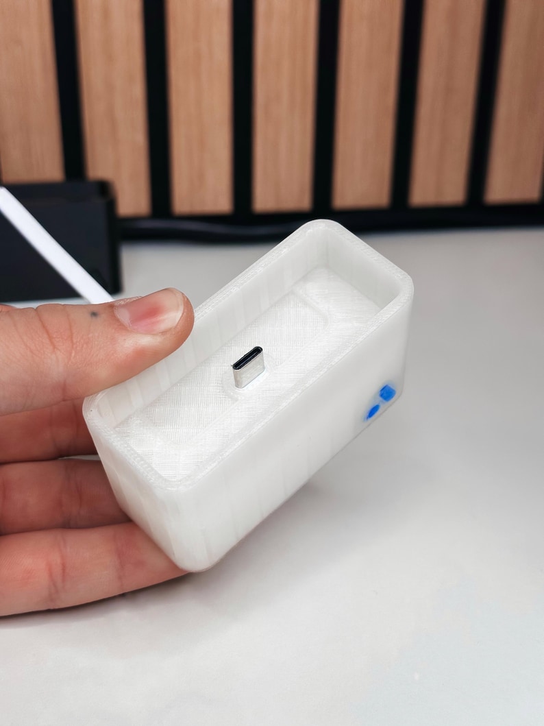 May include: A white rectangular charging dock with a USB-C port. The dock has a textured surface and blue accents on the side. The item is held in a hand.