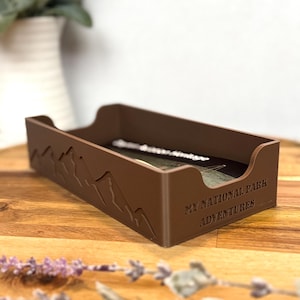 May include: A brown rectangular desk organizer with a mountain range design on the side. The text "MY NATIONAL PARK ADVENTURES" is printed on the side. The organizer is on a wooden surface.