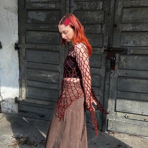 May include: A long-sleeved, dark red crochet net top with an open-weave pattern, worn over a black crop top, paired with a brown, ankle-length, paneled denim skirt and black platform boots, featuring a red hair clip in vibrant red hair.