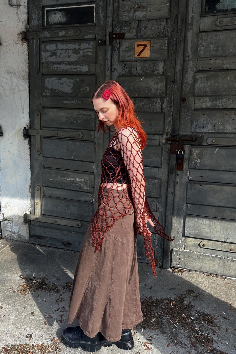 May include: A long-sleeved, dark red crochet net top with an open-weave pattern, worn over a black crop top, paired with a brown, ankle-length, paneled denim skirt and black platform boots, featuring a red hair clip in vibrant red hair.
