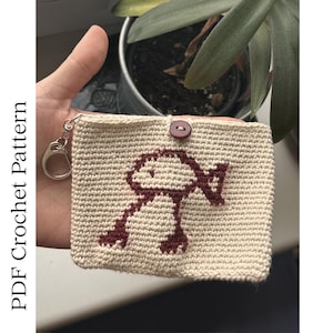 May include: A crocheted beige wallet with a maroon mushroom design. The wallet has a button closure and a silver key ring attached. The text "PDF Crochet Pattern" is printed on the left side of the image.