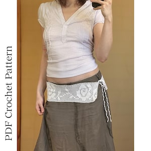 May include: A white crochet hip scarf featuring a detailed floral rose pattern with a side tie closure. The accessory is worn over a long, solid brown skirt and a light-colored short-sleeved top. The text PDF Crochet Pattern is visible.