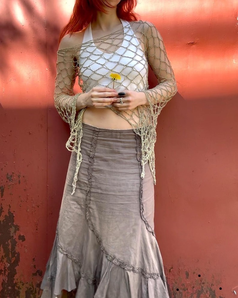 May include: A beige long-sleeve crochet net top with an open-weave pattern worn over a white halter-neck tank top, paired with a taupe midi skirt featuring vertical ruffled seams and a flared hemline that reaches below the knees.