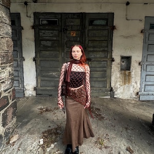 May include: A dark red long-sleeved fishnet top worn over a black tube top, paired with a brown maxi skirt. The outfit includes a matching dark red scarf draped around the neck and a dark brown shoulder bag, completed with black platform shoes.