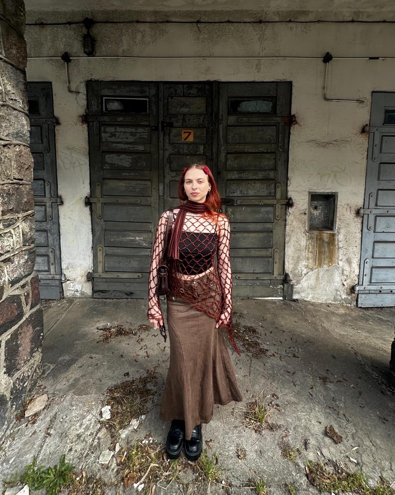 May include: A dark red long-sleeved fishnet top worn over a black tube top, paired with a brown maxi skirt. The outfit includes a matching dark red scarf draped around the neck and a dark brown shoulder bag, completed with black platform shoes.