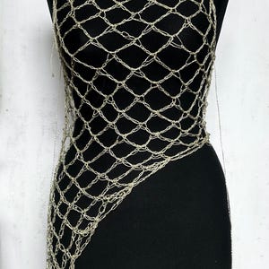 May include: A metallic silver crochet top featuring an open, diamond-shaped net pattern. The garment has a sleeveless design with a wide neckline and an asymmetrical hemline that extends longer on one side, displayed on a black mannequin.