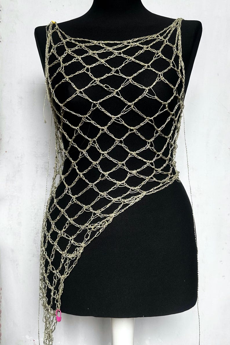 May include: A metallic silver crochet top featuring an open, diamond-shaped net pattern. The garment has a sleeveless design with a wide neckline and an asymmetrical hemline that extends longer on one side, displayed on a black mannequin.