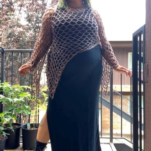 May include: A long-sleeved brown crochet net top with an asymmetrical hem worn over a solid black sleeveless maxi dress with a side slit, paired with black lace-up combat boots and black socks, creating a layered outfit with contrasting textures.