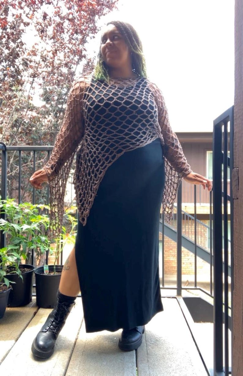 May include: A long-sleeved brown crochet net top with an asymmetrical hem worn over a solid black sleeveless maxi dress with a side slit, paired with black lace-up combat boots and black socks, creating a layered outfit with contrasting textures.