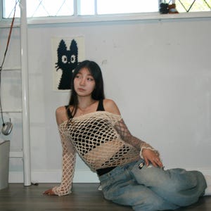 May include: A beige long-sleeve crochet fishnet top with an off-the-shoulder neckline worn over a black tank top, paired with light blue denim jeans. The top features a wide-gauge open knit pattern that extends down to the wrists.