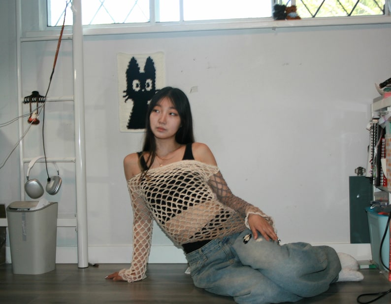 May include: A beige long-sleeve crochet fishnet top with an off-the-shoulder neckline worn over a black tank top, paired with light blue denim jeans. The top features a wide-gauge open knit pattern that extends down to the wrists.