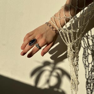 May include: A cream-colored crocheted net sleeve featuring an open-weave pattern. The wrist is adorned with a silver-toned chain link bracelet. The ring finger displays a silver ring with an oval black stone, and the index finger has a silver ring.