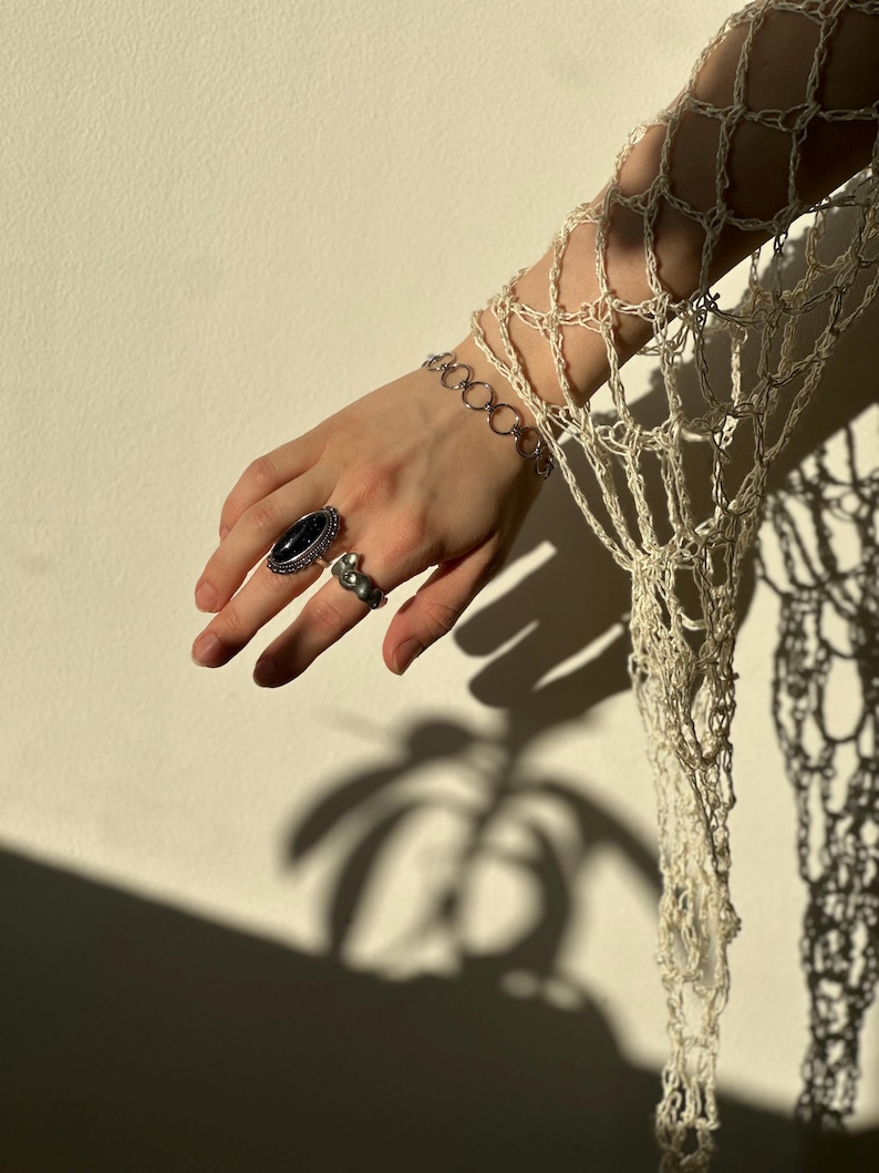 May include: A cream-colored crocheted net sleeve featuring an open-weave pattern. The wrist is adorned with a silver-toned chain link bracelet. The ring finger displays a silver ring with an oval black stone, and the index finger has a silver ring.