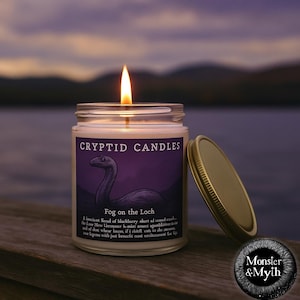 May include: A purple and gold candle jar with a label that reads "CRYPTID CANDLES" and "Fog on the Loch". The candle is lit and the jar has a gold lid. The label features a black and white illustration of a Loch Ness Monster. The candle is on a wooden surface with a blurred background of a lake and sky.