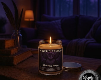 May include: A lit candle in a glass jar with a black and purple label that reads "Cryptid Candles" and "When Wings Whisper". The candle is on a wooden table with a stack of books and a sofa in the background.