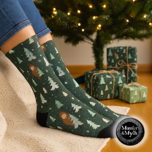 May include: Dark green socks with a pattern of white and green Christmas trees and a cartoon Bigfoot. The socks have black heels and toes. The socks are near wrapped gifts and a Christmas tree.
