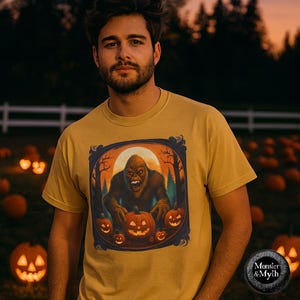 Peut inclure: T-shirt jaune moutarde avec un motif de Bigfoot entouré de citrouilles d'Halloween et d'arbres, sur fond de pleine lune. Le t-shirt a un col rond et des manches courtes. Le motif est encadré d'une bordure bleu foncé. Collection Monster & Myth.