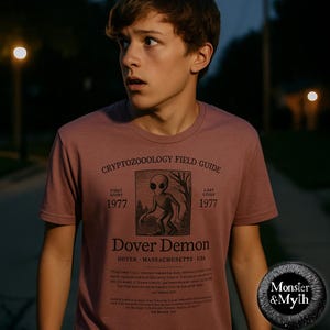 May include: A person wearing a maroon t-shirt with a black and white graphic of a creature from the Dover Demon legend. The graphic includes the text "Cryptozoology Field Guide", "First Sight 1977", "Last Sight 1977", "Dover Demon", "Dover, Massachusetts, USA", and a description of the creature. The t-shirt also has the text "Monster & Myth" on the bottom right corner.