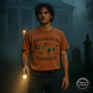 Op de afbeelding: Een oranje t-shirt met de woorden "PARANORMAL TENNESSEE" op de voorkant gedrukt. Het shirt heeft verschillende kleine afbeeldingen. De persoon op de afbeelding houdt een zaklamp vast. De achtergrond omvat een spookachtig gebouw en een mistige sfeer.