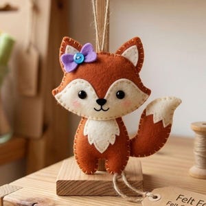 Könnte beinhalten: Handgefertigter Fuchsschmuck aus Filz, braun mit weißen Akzenten und einer lila Schleife. Er hängt an einer Schnur. Ein Etikett lautet "Felt Fox Felt Ornament - Handmade -."
