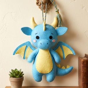 May include: A handmade blue felt dragon with yellow wings, belly, and horns hangs from a decorative wall hook. The dragon has a friendly expression with pink cheeks and is displayed on a wooden shelf with a small potted plant and antique books.