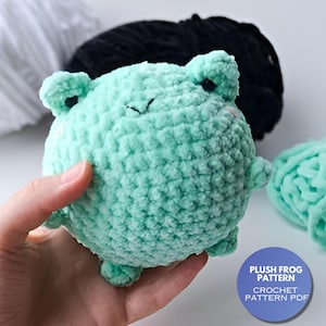 May include: A crocheted plush frog toy in a light green color. The frog has a round body and small legs. The toy is being held in a hand. The text "PLUSH FROG PATTERN CROCHET PATTERN PDF" is on a blue circle in the lower right corner of the image.